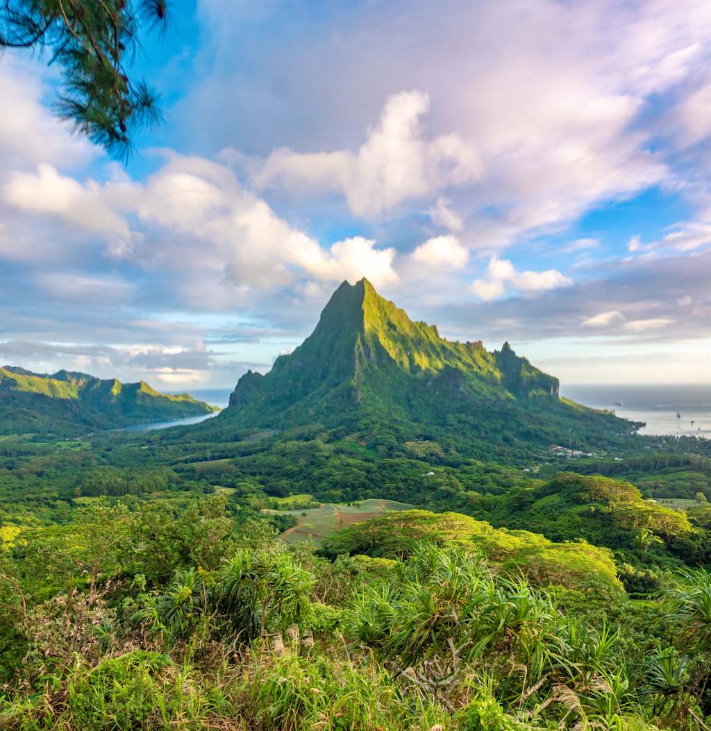 A lush, verdant landscape with a towering, rugged mountain peak rising in the distance, surrounded by a dramatic cloudy sky.