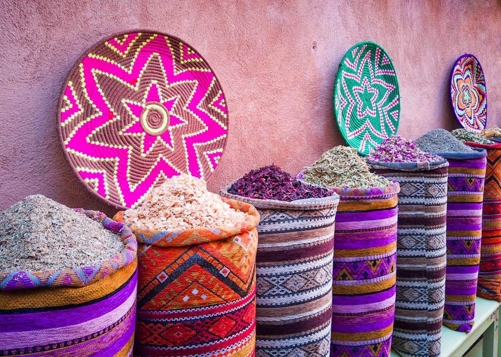 Vibrant, patterned baskets and decorative items in various shades of pink, purple, and green are displayed against a textured, colorful wall.