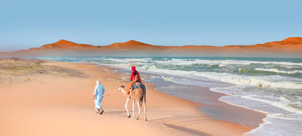 Two people in colorful clothing walking along the sandy beach, with towering orange mountains in the background and a calm, blue ocean.