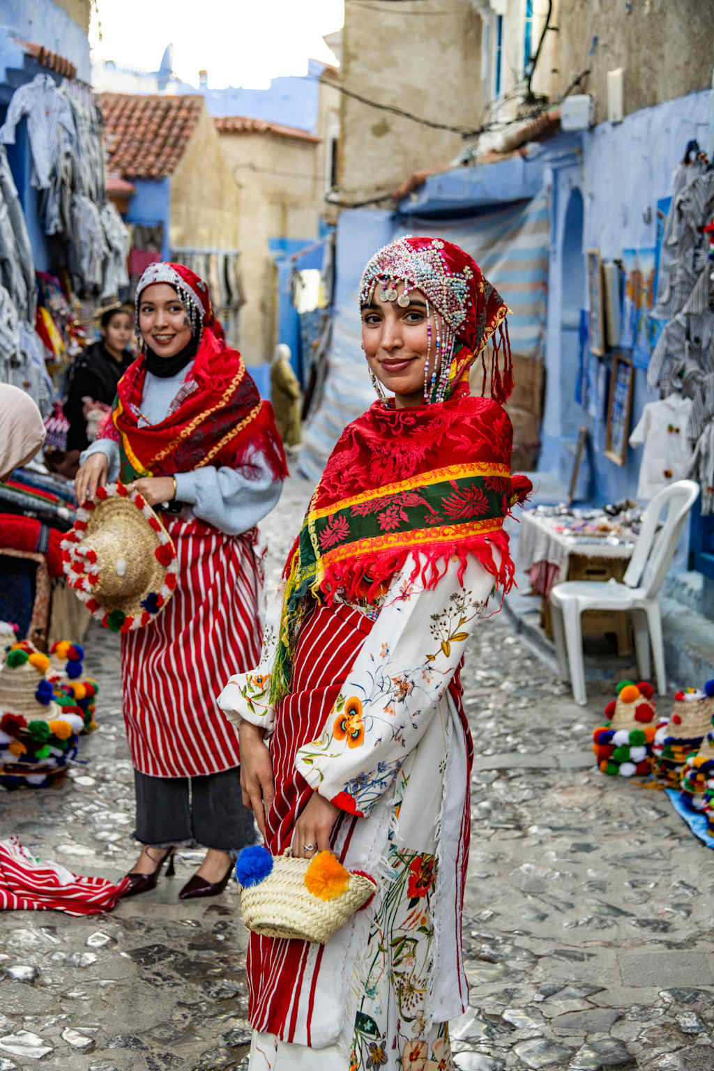 Two women in traditional Moroccan clothing, with vibrant red and white patterns, stand in a narrow alley lined with colorful buildings and merchandise.