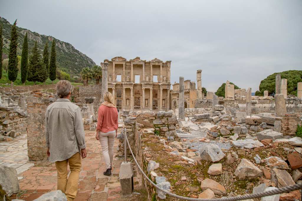 The image shows two people walking through the ancient ruins of the Library of Celsus, a well-preserved Roman structure in the historic city of Ephesus, surrounded by the remnants of other structures and a mountainous landscape.