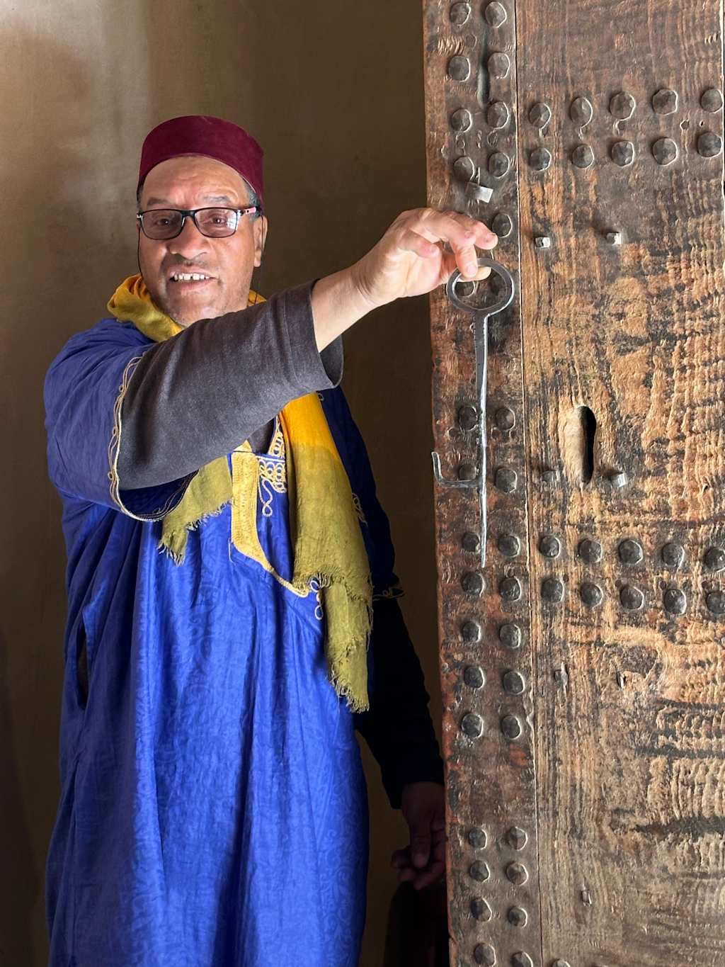 A man in traditional blue clothing and a red fez uses a large key to unlock a rustic wooden door, with a warm, earthy background.