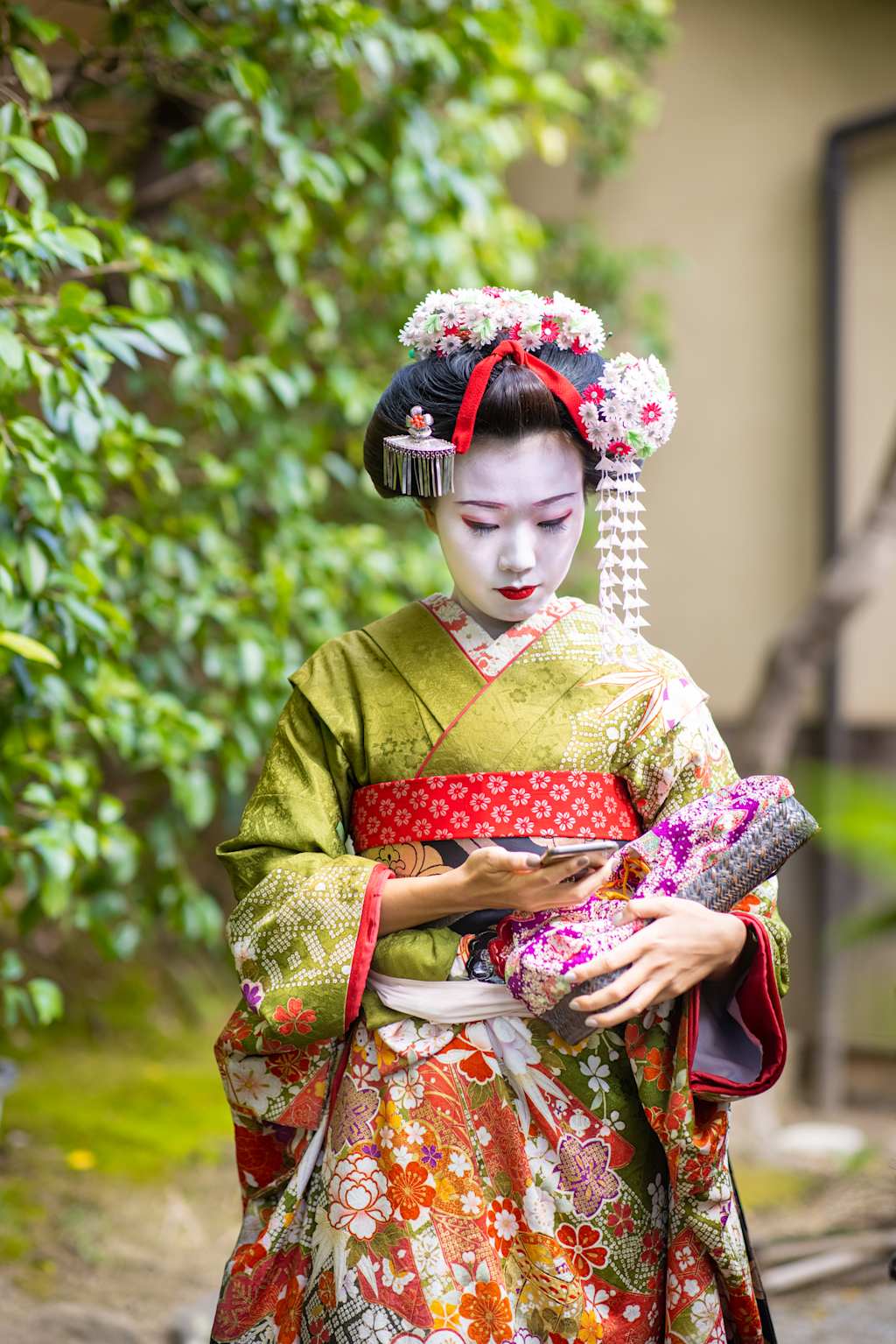 A geisha in a vibrant kimono holds a patterned purse while standing in front of lush green foliage.