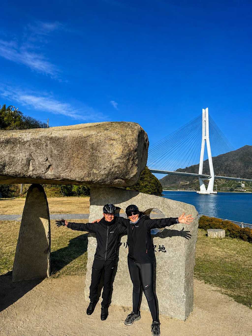 Two people in cycling gear pose with outstretched arms in front of a large stone structure, with a white suspension bridge spanning a blue body of water in the background under a clear sky.