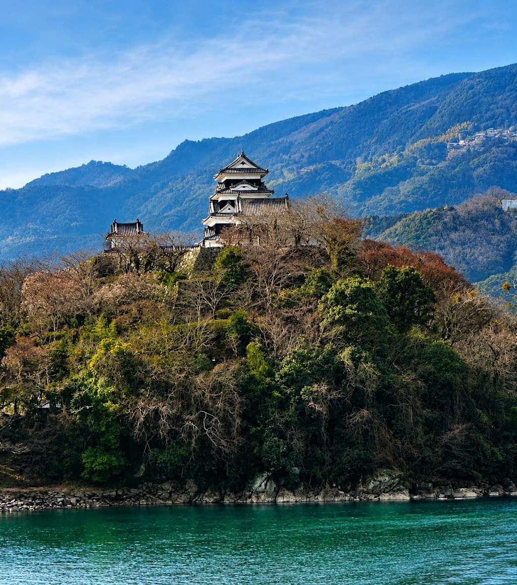 A Japanese castle stands atop a tree-covered hill in the foreground, with lush, blue mountains rising in the background under a clear sky.