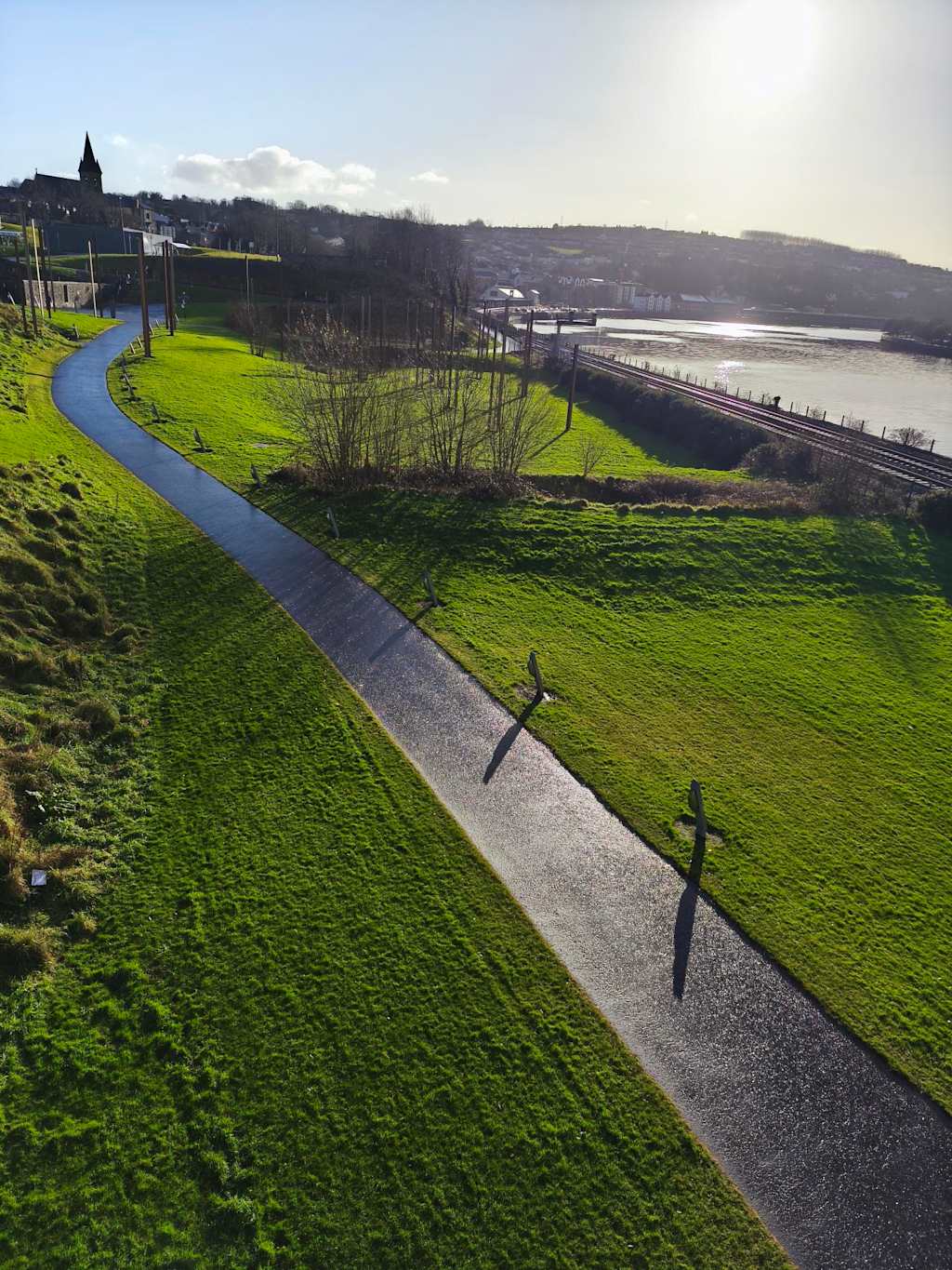 A paved path winds through a grassy hillside in the foreground, leading towards a river and distant town under a bright, hazy sun in the background.