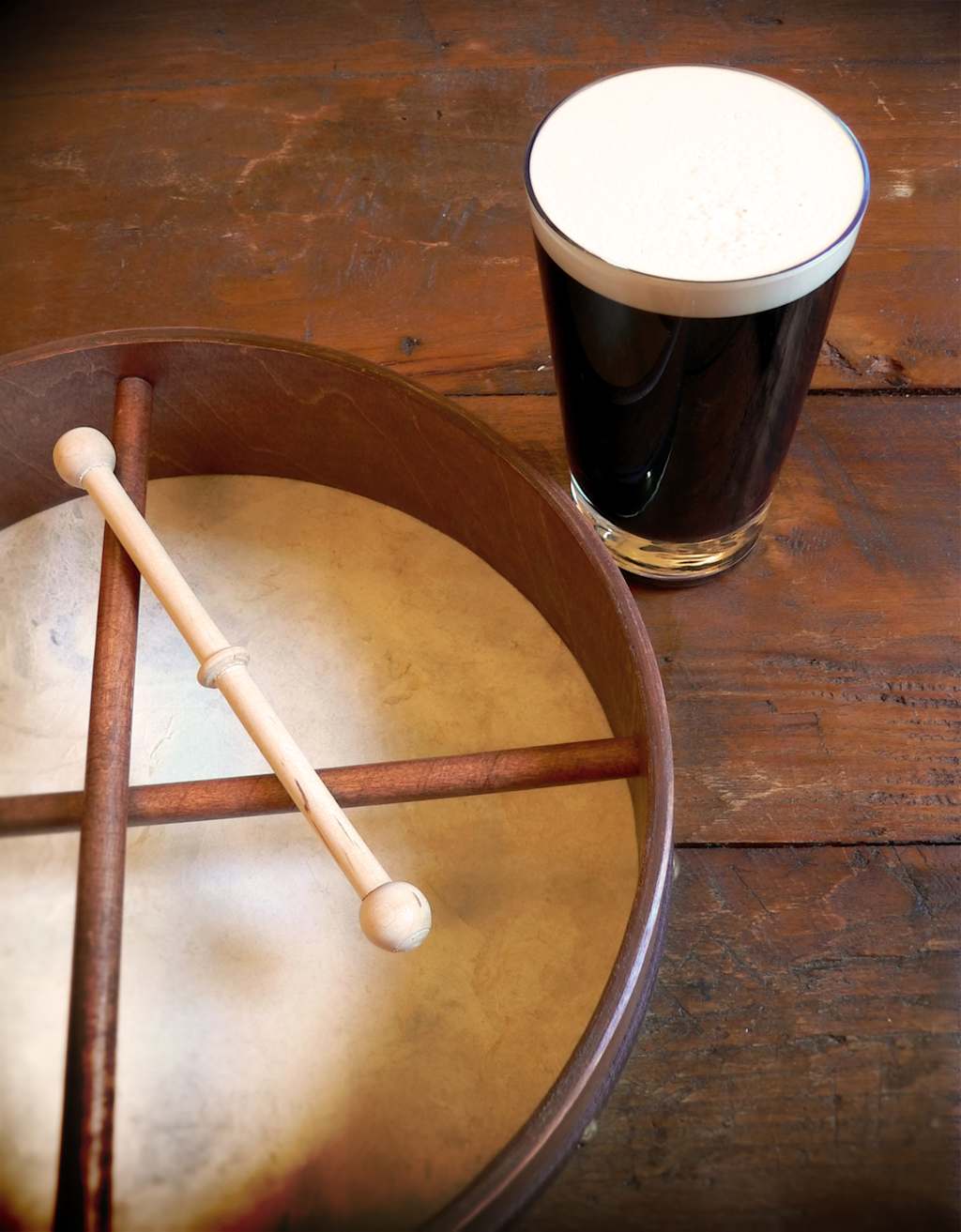 A bodhrán with a beater rests on a wooden surface next to a pint of dark beer with a frothy head.