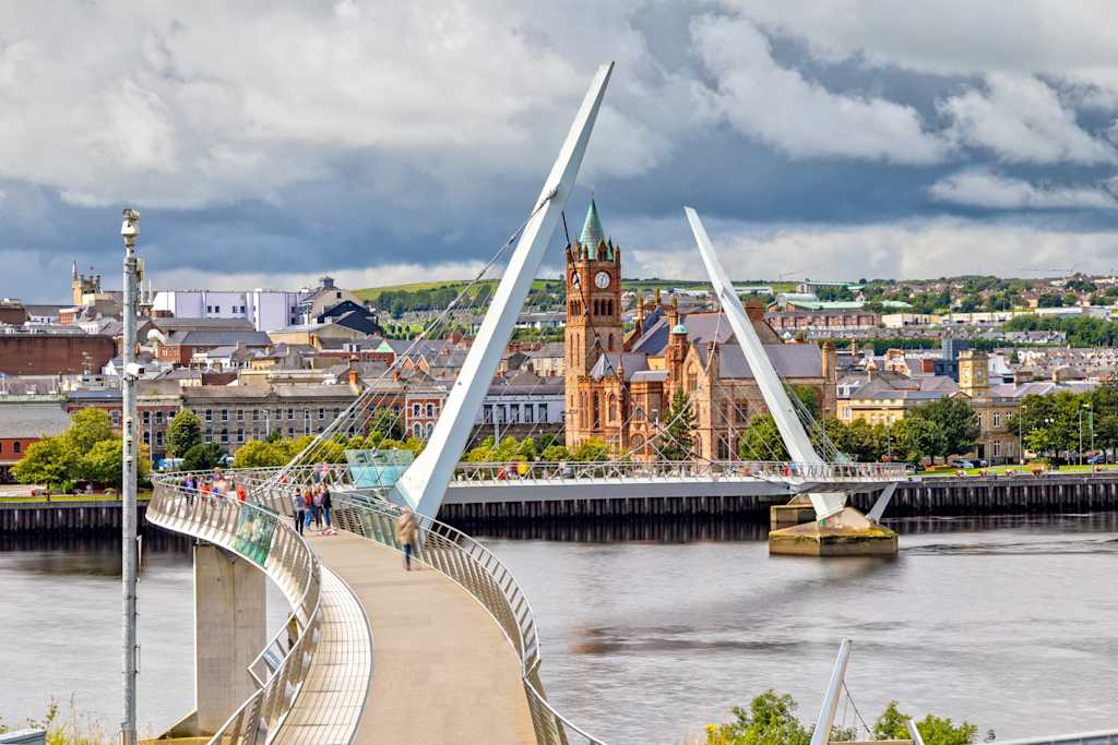 A modern pedestrian bridge curves across a river in the foreground, leading to a city skyline with a prominent clock tower in the background under a cloudy sky.