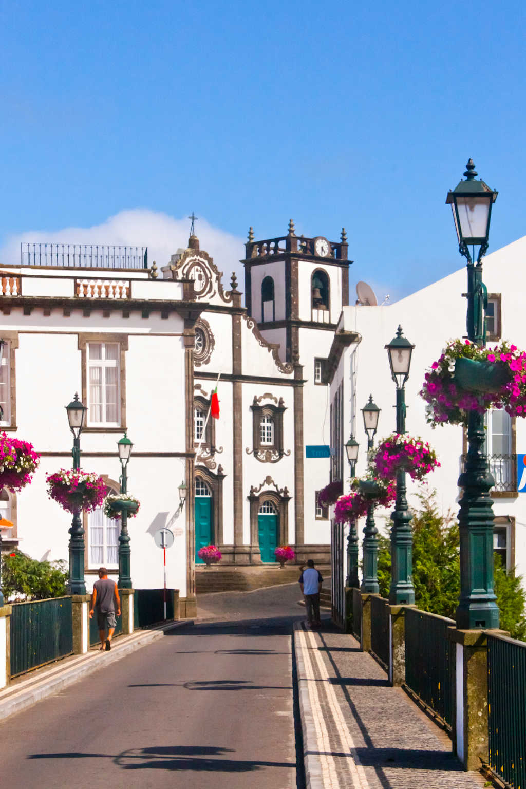 A bridge with lampposts adorned with pink flowers leads to a white building with a bell tower under a clear blue sky.