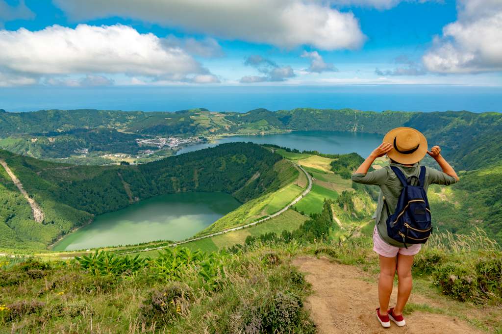 A hiker with a backpack and hat stands on a grassy overlook, gazing at a vast caldera with two lakes and a distant ocean under a cloudy blue sky.