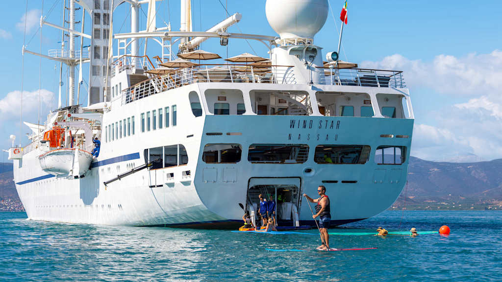 A large white cruise ship with various antennas and equipment on its deck is anchored in a body of water, surrounded by people swimming and enjoying the sunny day.