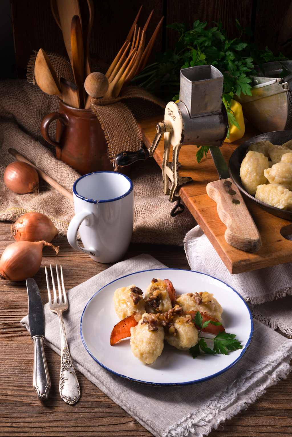 A plate of dumplings with bacon and parsley sits on a napkin in the foreground, while a rustic kitchen scene with utensils and ingredients fills the background.