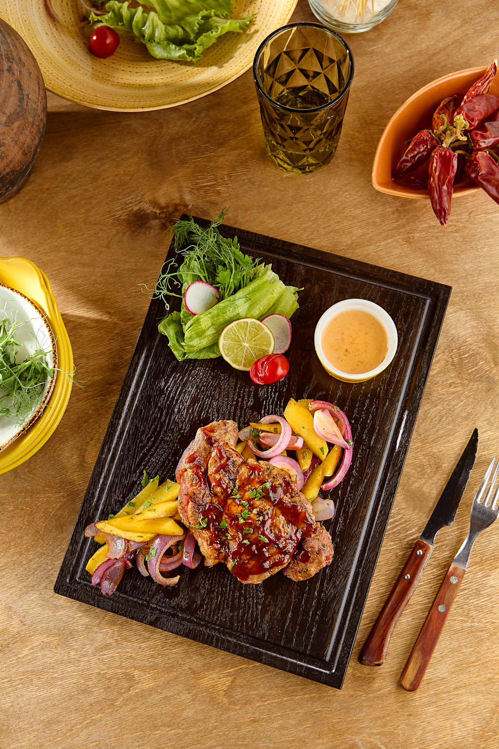 A dark wooden platter holds a steak with mango and red onion salsa, a small dish of sauce, and a side salad, all set on a rustic wooden table with a glass of water and a bowl of crawfish in the background.