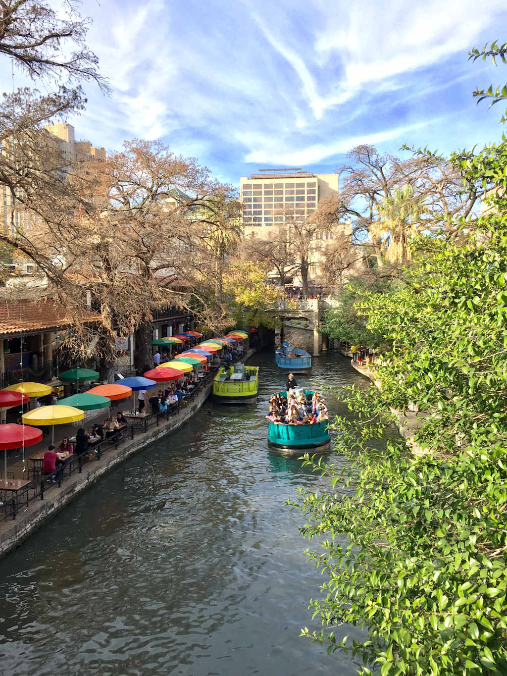 Colorful umbrellas shade diners along a riverwalk where tour boats navigate the water, with buildings and trees forming the backdrop under a cloudy sky.