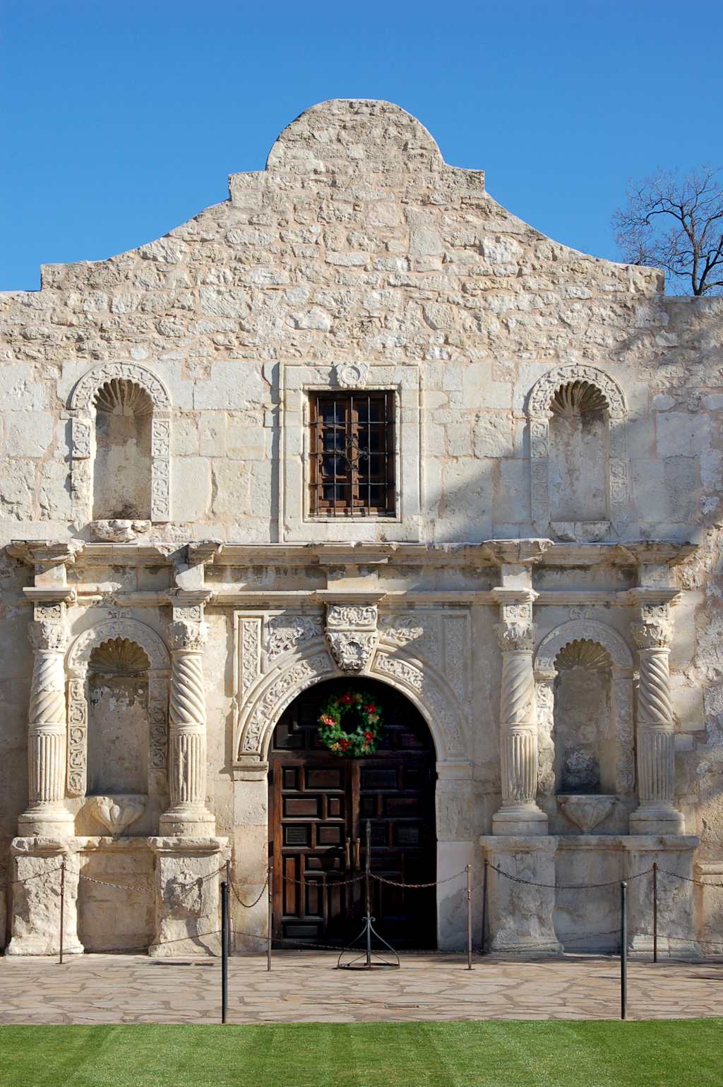 A weathered stone facade with a wooden door adorned with a wreath stands against a clear blue sky, with manicured green grass in the foreground.
