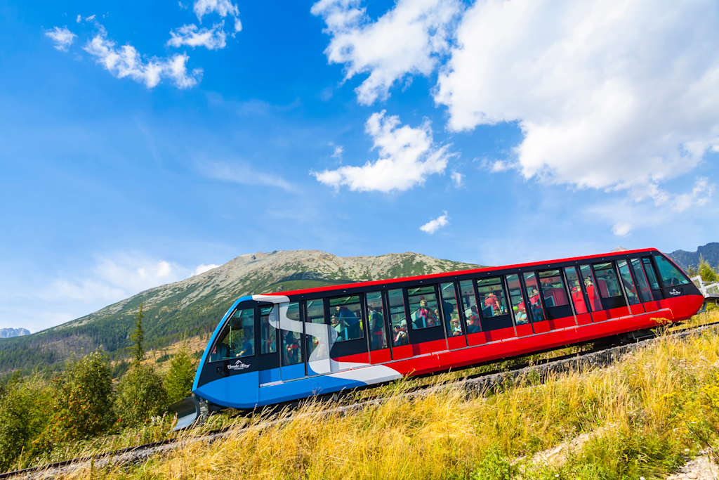 A red and blue funicular train ascends a grassy slope with a mountain range and a bright blue sky with clouds in the background.