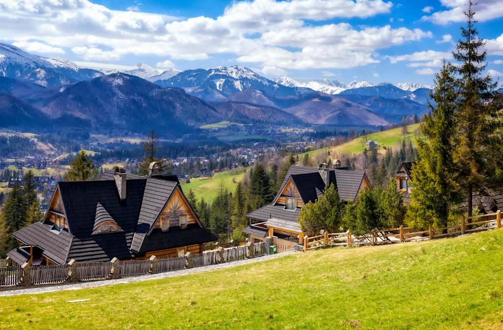 Two wooden houses with dark roofs sit on a grassy hill in the foreground, with a valley town and snow-capped mountains in the background.