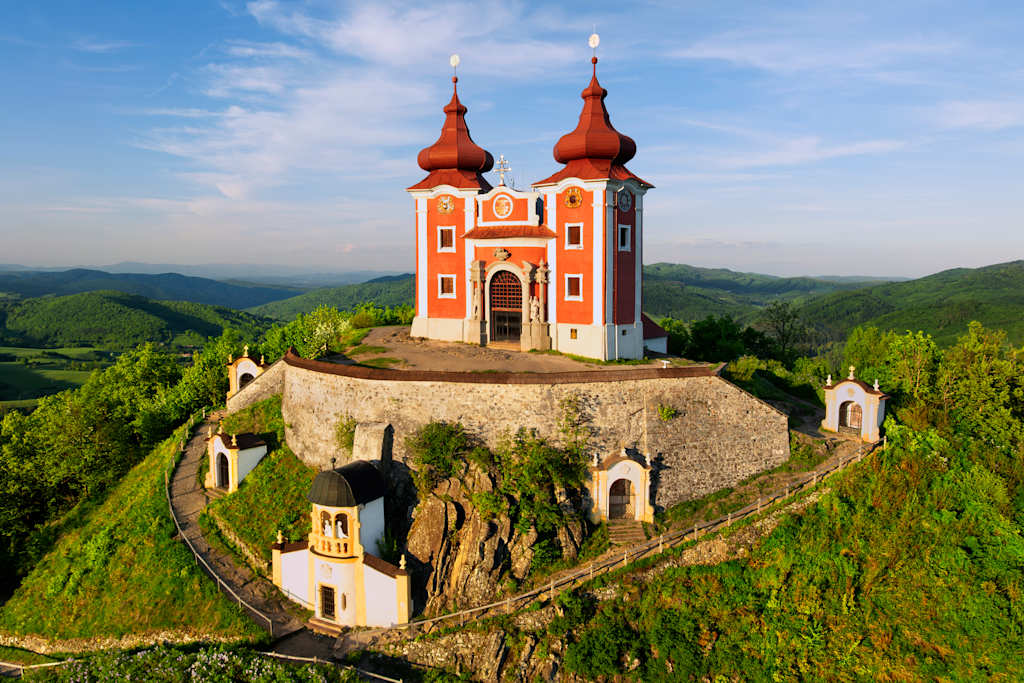 A striking red and white church with twin onion domes sits atop a stone-walled hill in the foreground, while rolling green hills and a hazy blue sky form the background.
