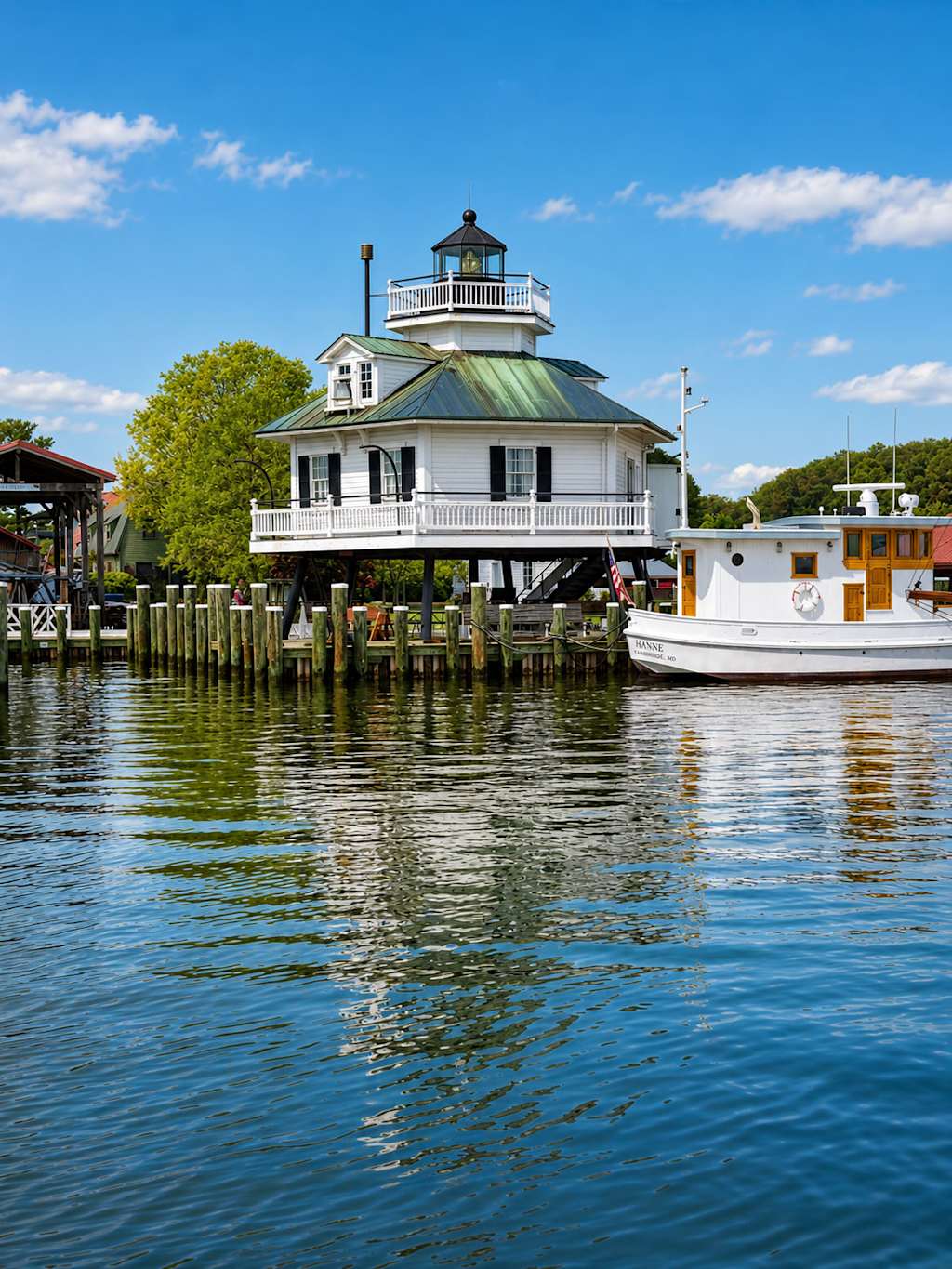 A white lighthouse on stilts stands over a wooden pier in the foreground, with a white boat docked beside it and lush green trees under a bright blue sky in the background.