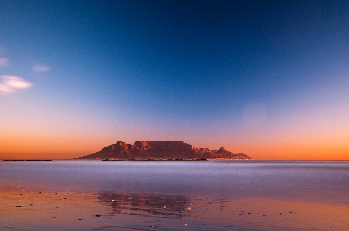 A serene landscape with a majestic mountain range silhouetted against a vibrant orange and blue sky, reflected in the calm waters of the foreground.