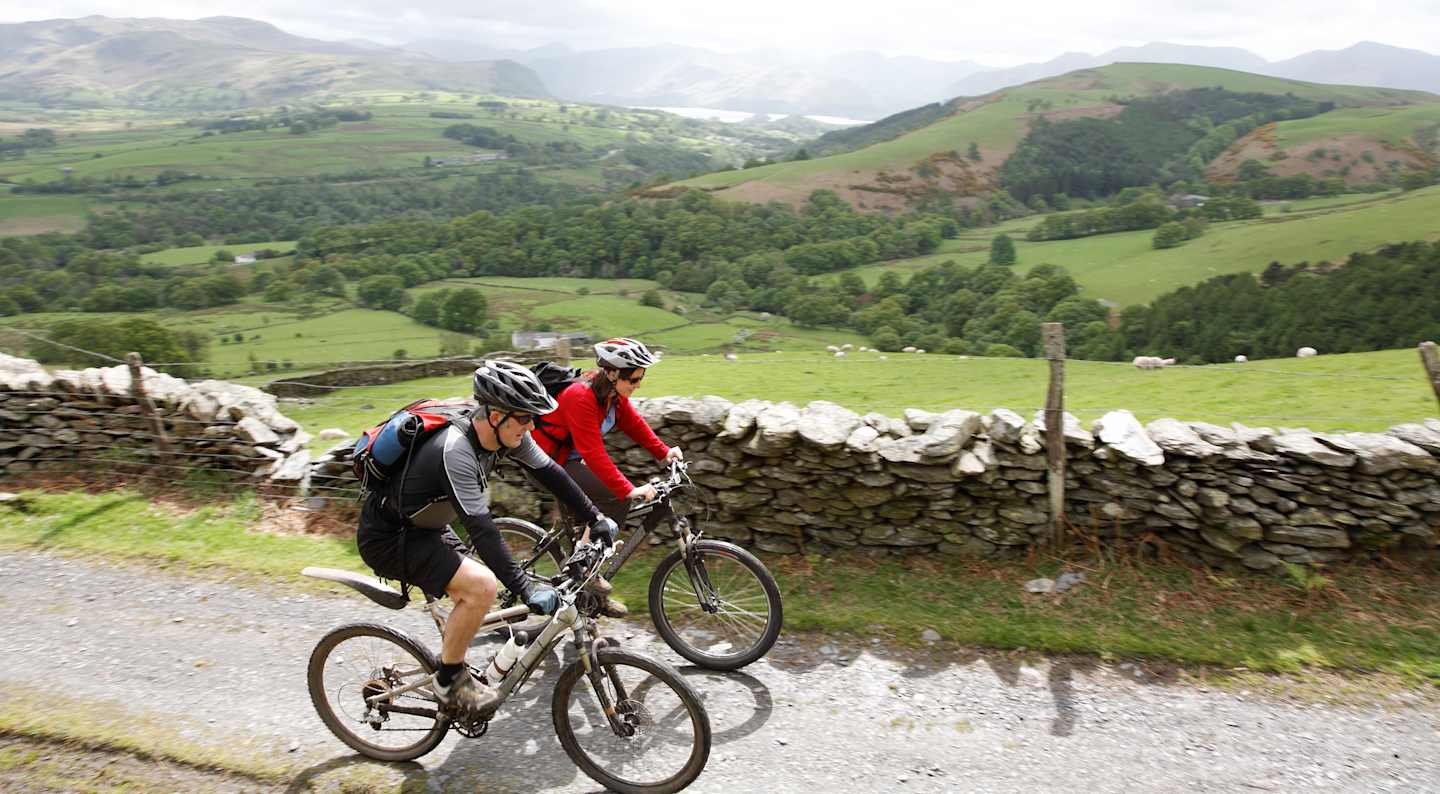 A cyclist rides along a dirt path surrounded by lush green hills, stone walls, and a picturesque rural landscape in the background.