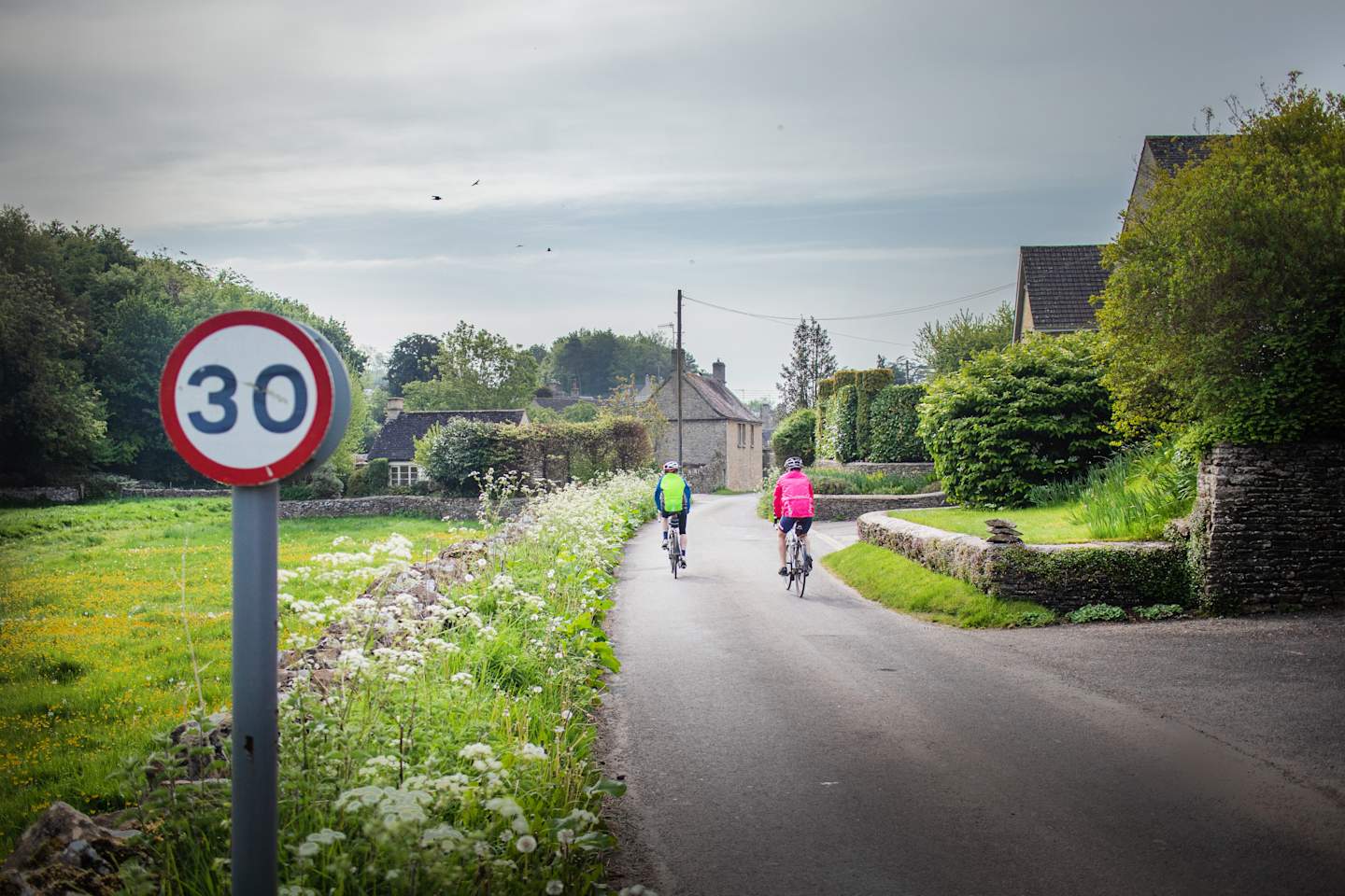 Two cyclists riding past a 30mph sign through the village of Daglingworth, near Cirencester, Gloucestershire. It is situated in the Duntisbourne valley. The name is derived from the river Dunt which flows through the village.