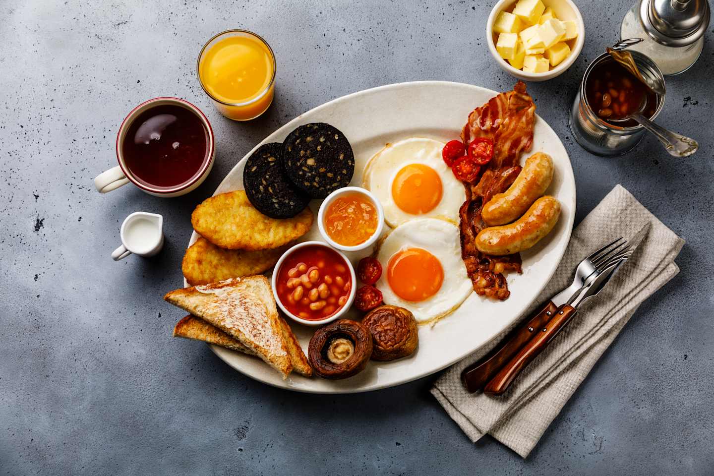 Full fry up English breakfast with fried eggs, sausages, bacon, black pudding, beans, toasts and tea on gray concrete background