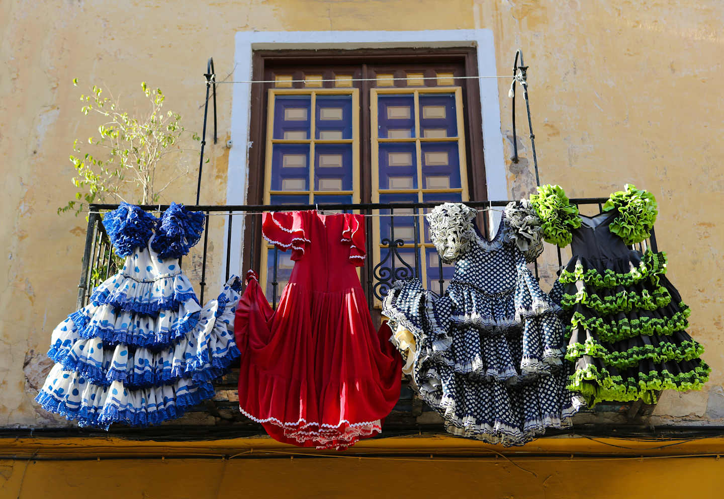 Colorful flamenco dresses hang on a balcony, with a yellow-walled building and a wooden window frame in the background.