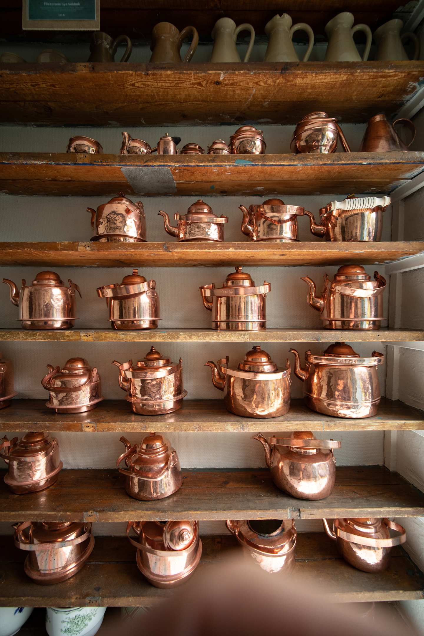 A collection of various copper cookware items, including pots, pans, and teapots, neatly arranged on wooden shelves against a dark background.