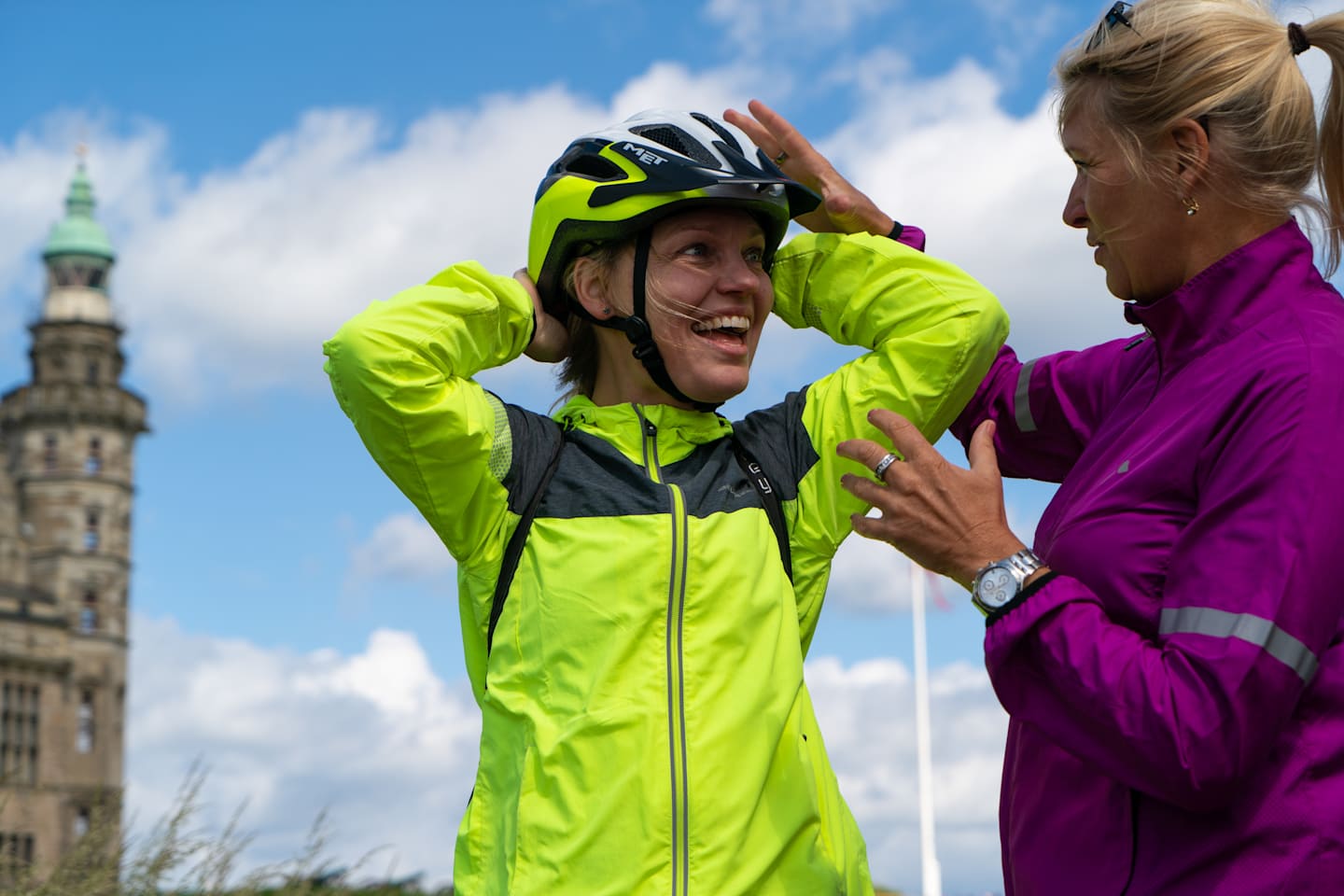 A woman in a bright yellow and black cycling jacket is adjusting her helmet, with a historic building visible in the background.