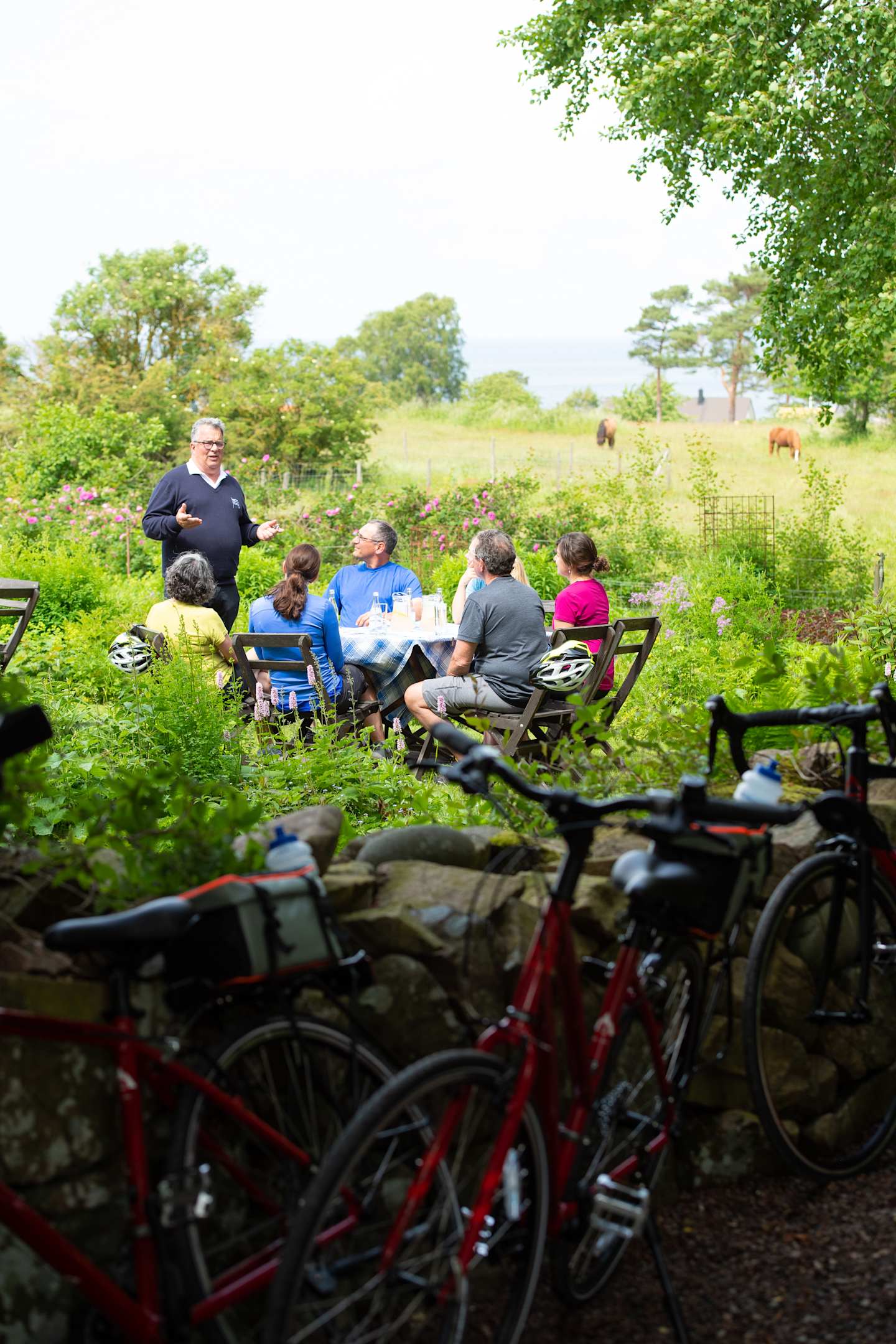 A group of people are gathered around a table in a scenic outdoor setting, with bicycles in the foreground and a pastoral landscape in the background.
