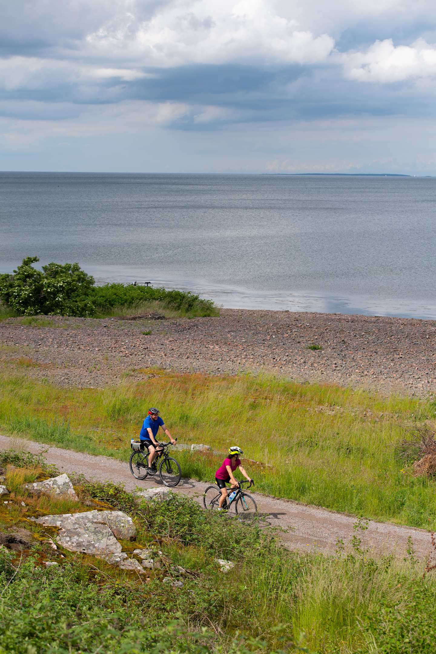 Two cyclists riding on a dirt path along the shoreline, with a vast body of water and cloudy sky in the background.