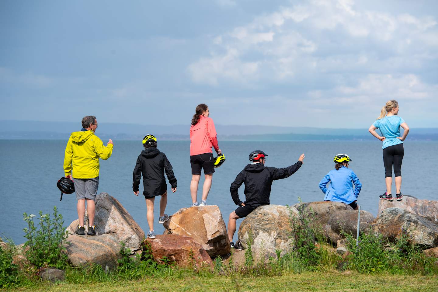A group of people in colorful outdoor gear standing on rocky outcroppings overlooking a scenic coastal landscape with mountains in the distance.