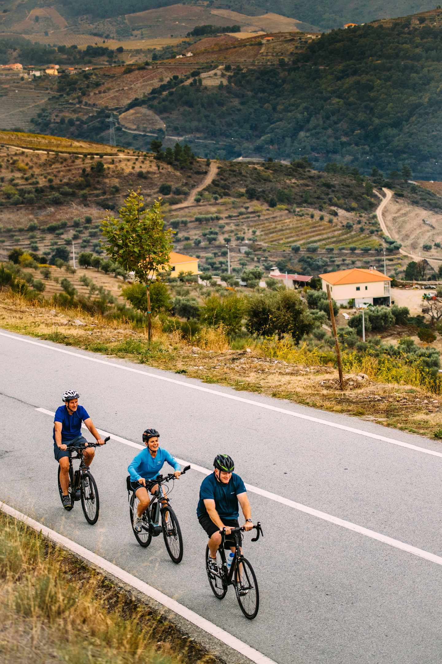 A group of cyclists riding on a winding road through a hilly, rural landscape with scattered buildings and vegetation in the background.