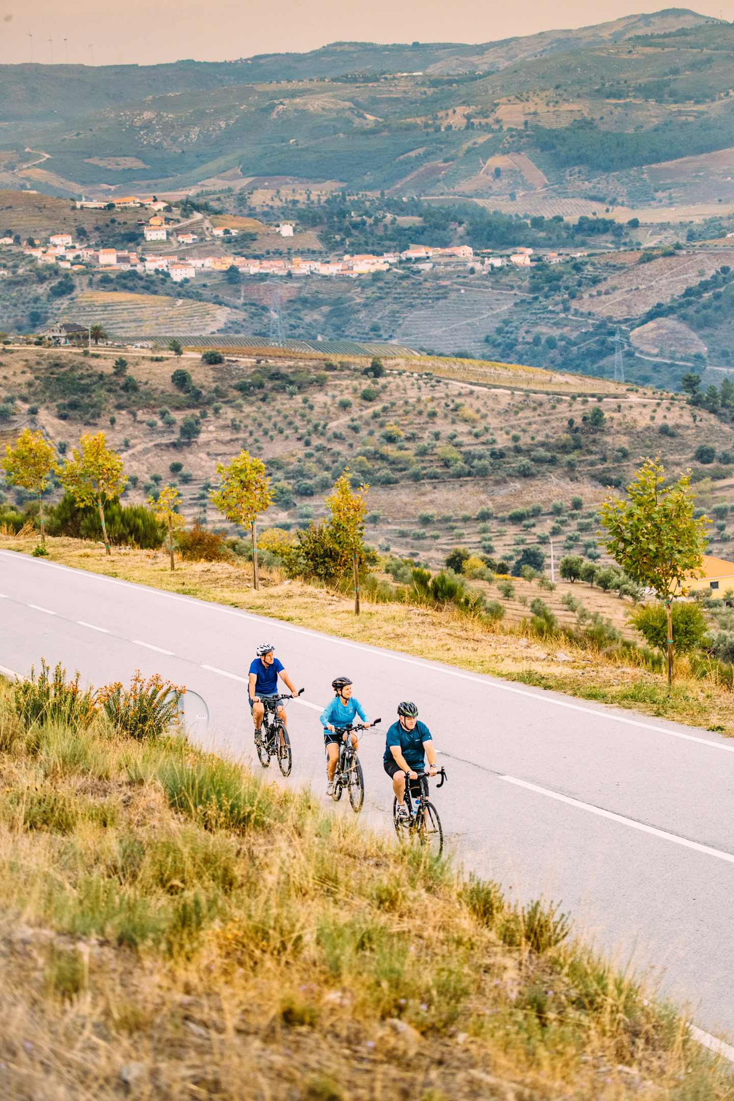 A group of cyclists riding on a winding road through a scenic landscape with rolling hills, distant villages, and a hazy blue sky in the background.