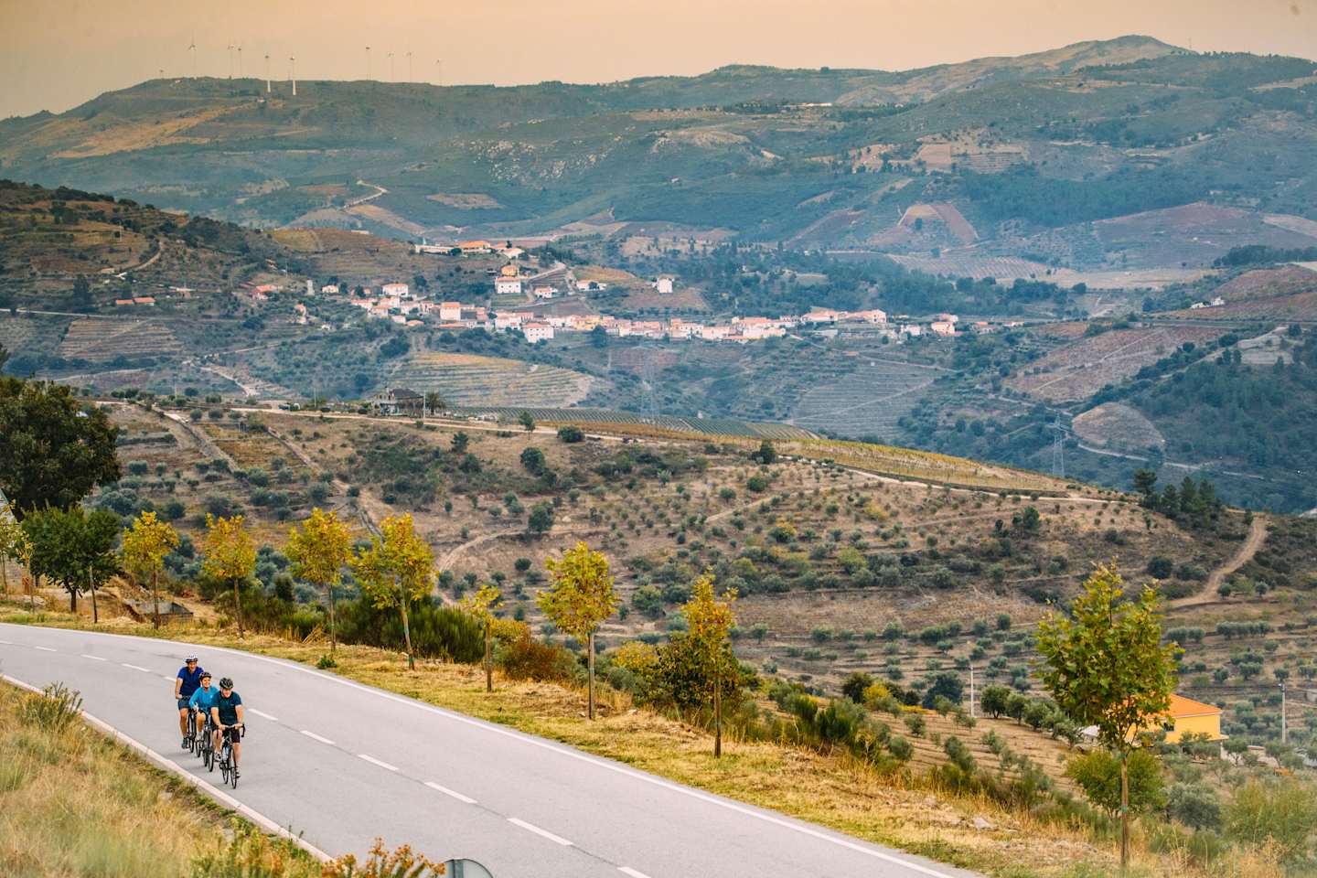 A winding road winds through a hilly, rural landscape with olive groves and small villages nestled in the valleys below the mountains in the distance.
