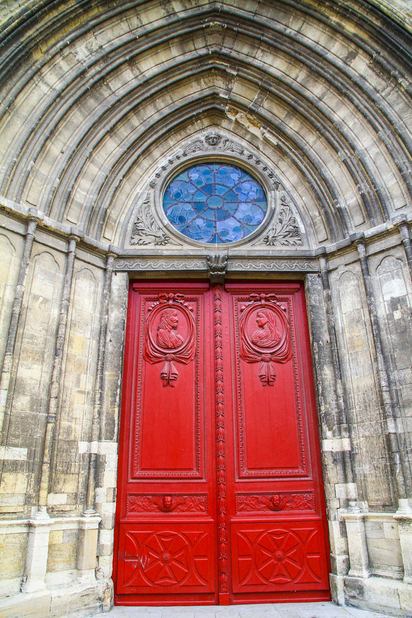 A grand, ornate archway with a stained glass window above a pair of striking red wooden doors, set against a backdrop of intricate stone architecture.