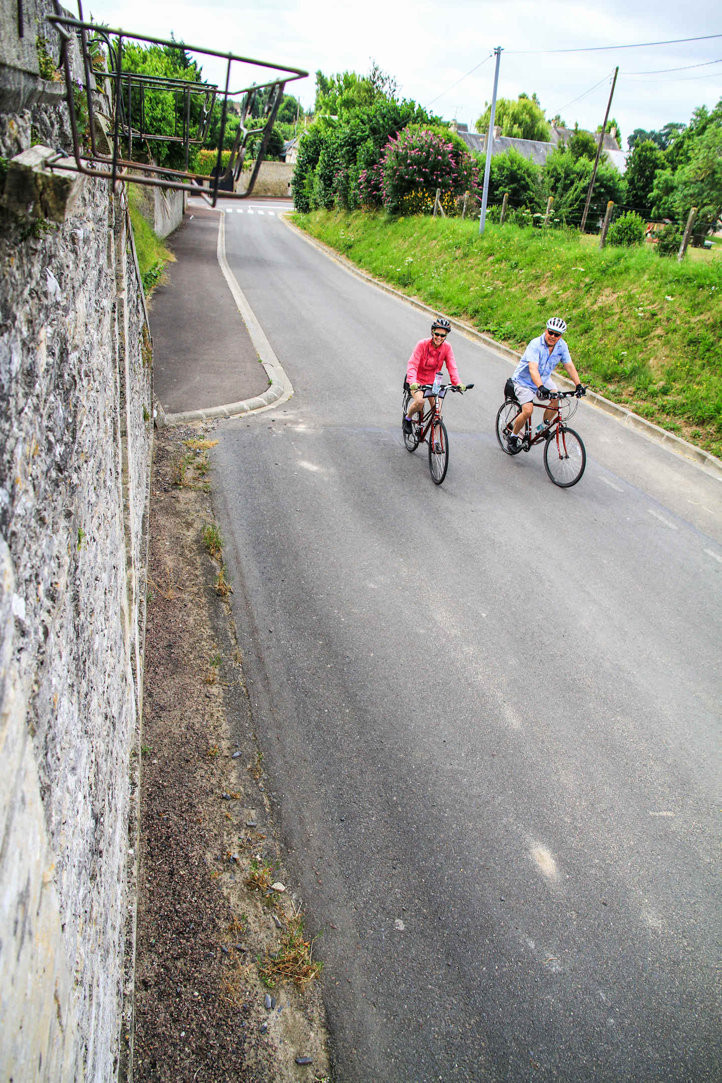 Two cyclists riding bicycles on a paved road surrounded by lush greenery and a stone wall in the background.