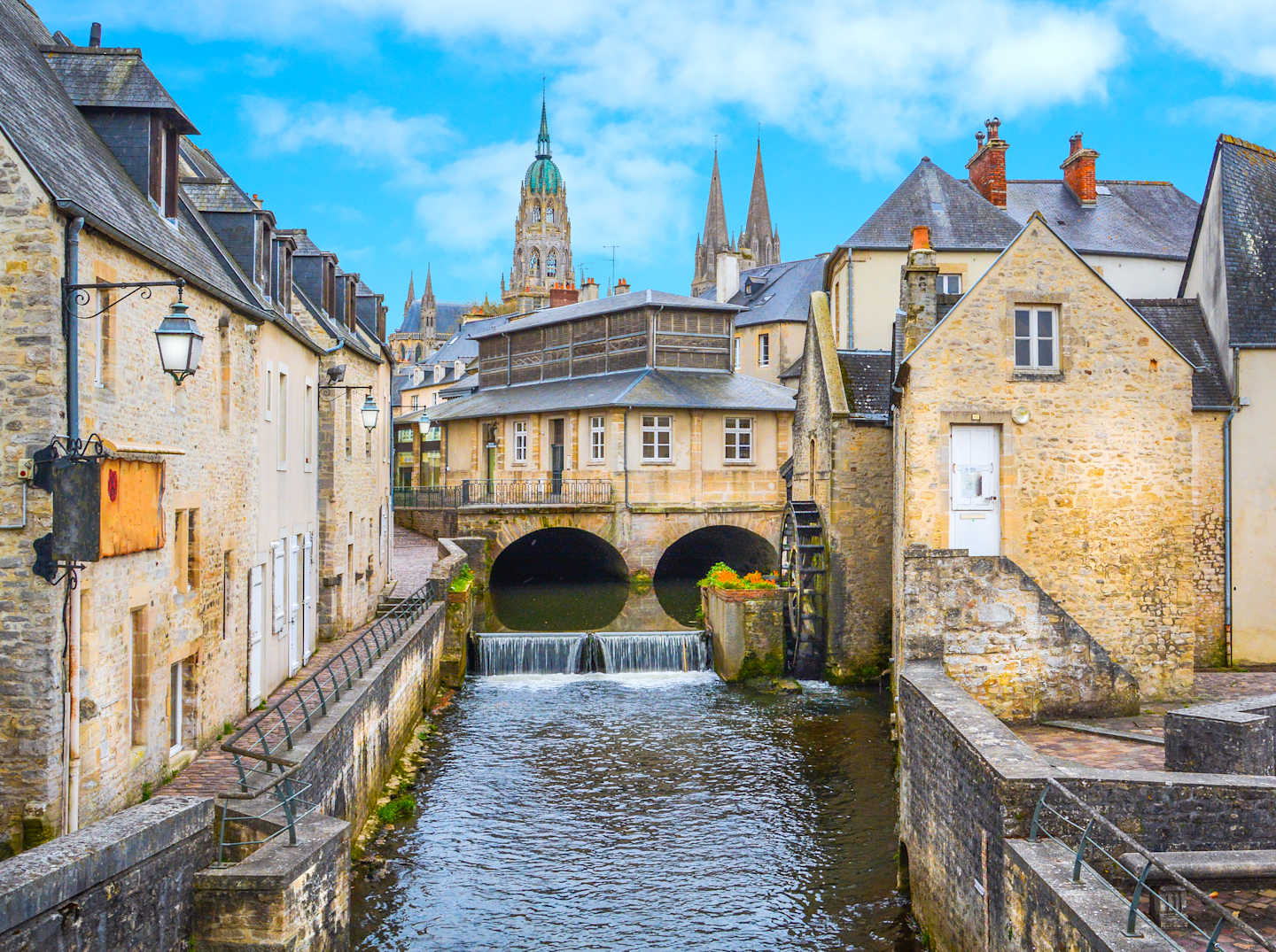 A picturesque canal winds through a historic town, with charming stone buildings, bridges, and church spires rising in the background against a cloudy blue sky.