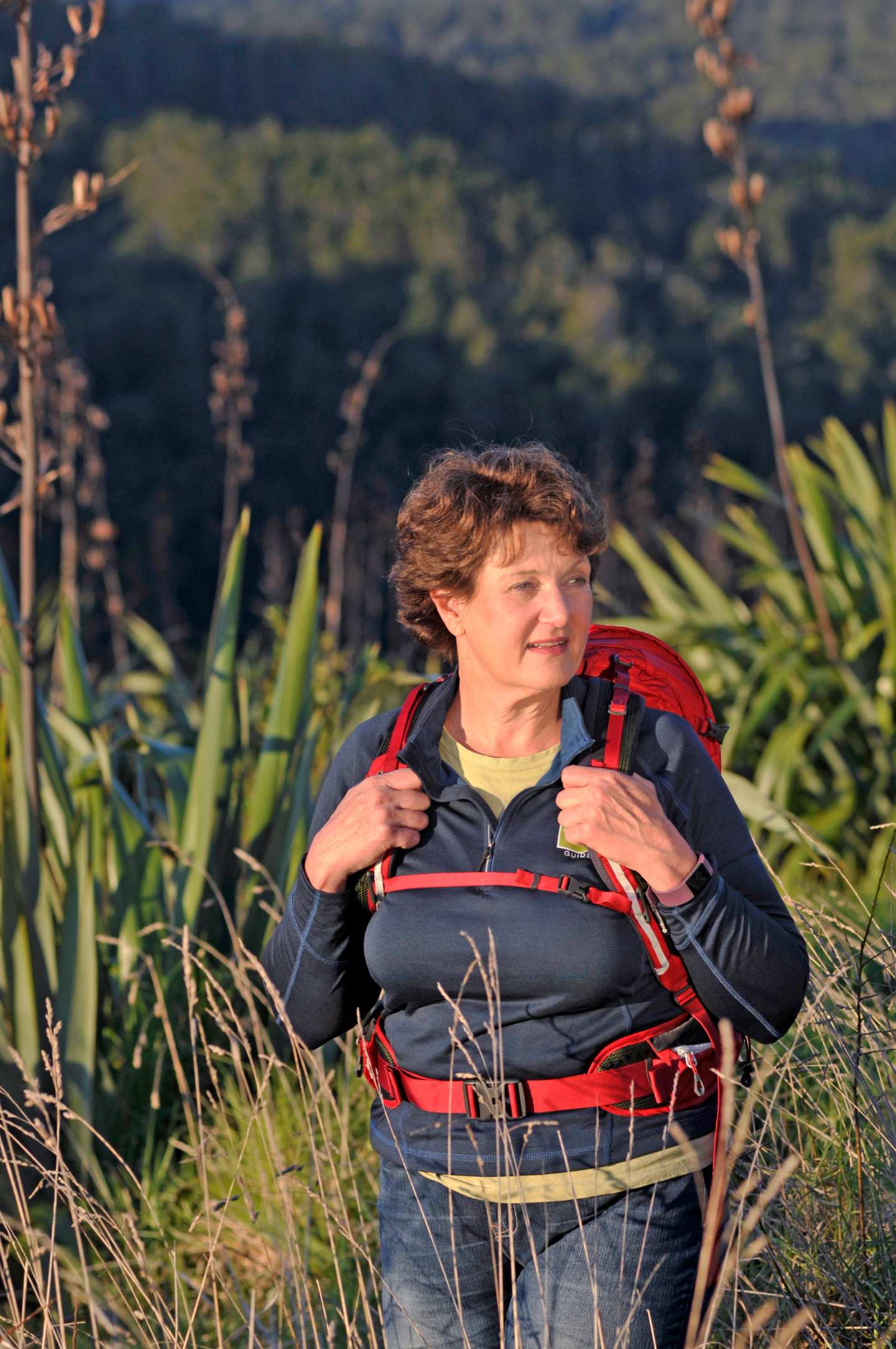 A person wearing a red and black backpack stands in a grassy field with a forested area in the background.