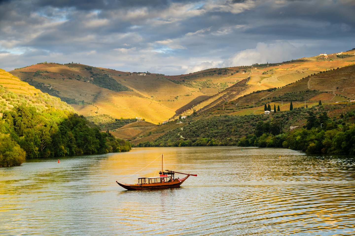 A serene river flows through a picturesque landscape of rolling hills, dotted with trees and a traditional boat sailing across the calm waters, set against a dramatic cloudy sky.