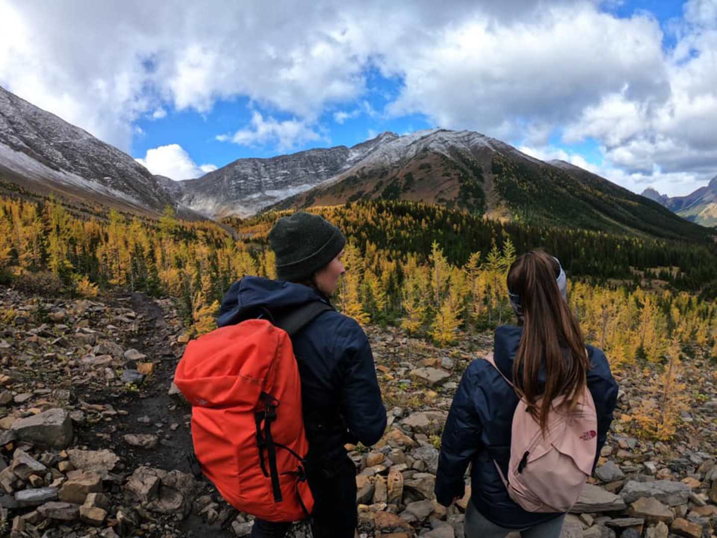 Two people in outdoor gear stand on a rocky path, gazing at the majestic mountains and autumn-colored forests in the background.