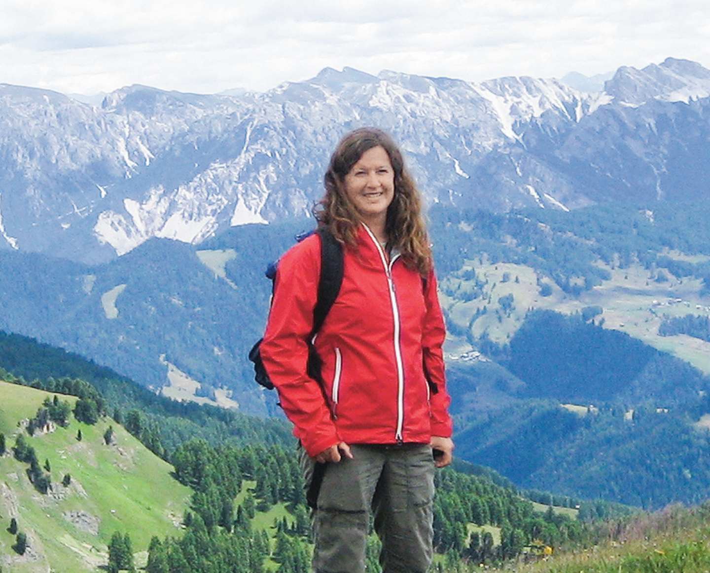 A woman in a red jacket stands in the foreground, with a breathtaking mountain landscape in the background.
