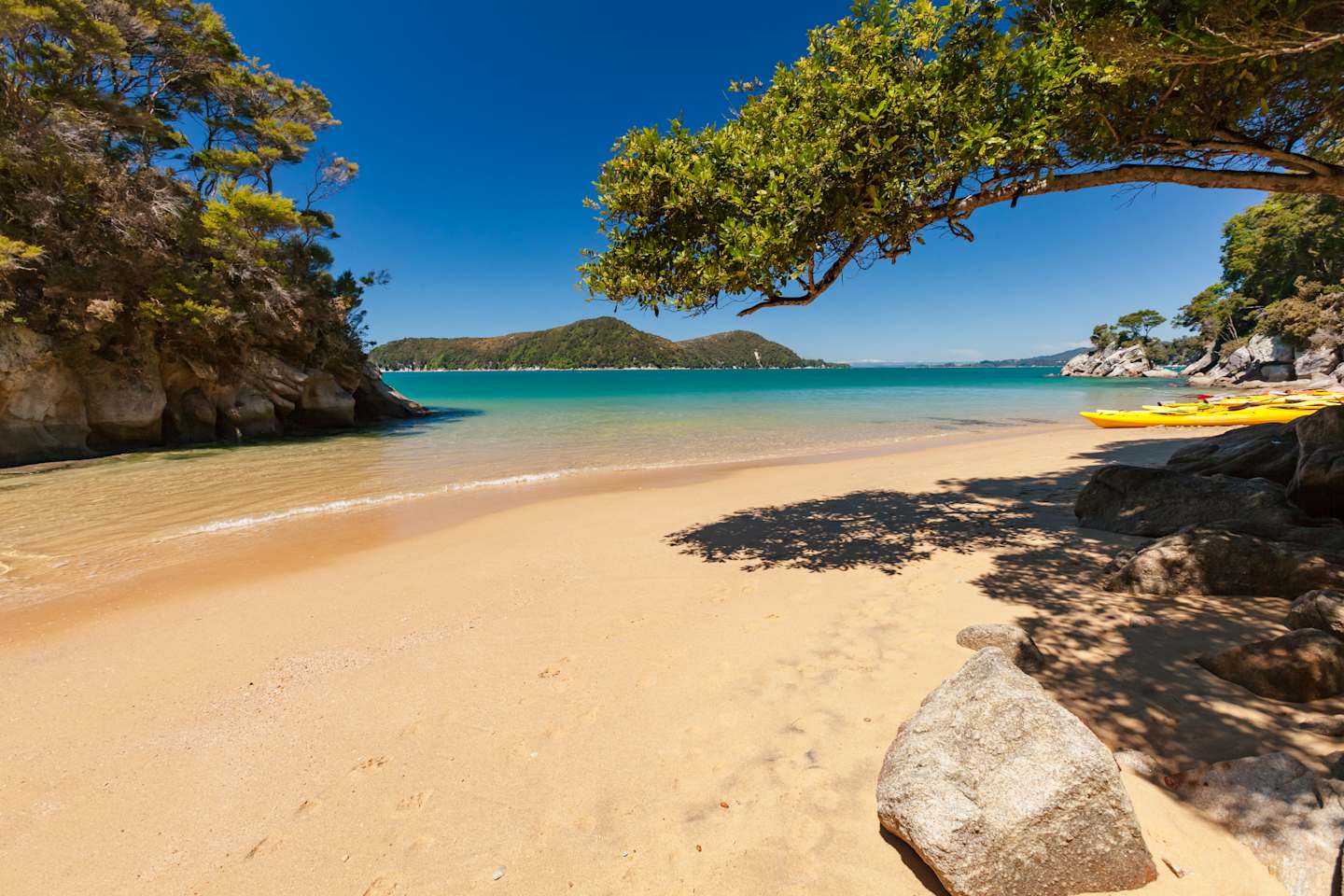 A serene beach with golden sand, surrounded by lush green trees and rocky cliffs, with a clear blue sky and calm turquoise waters in the background, and several yellow kayaks resting on the shore.