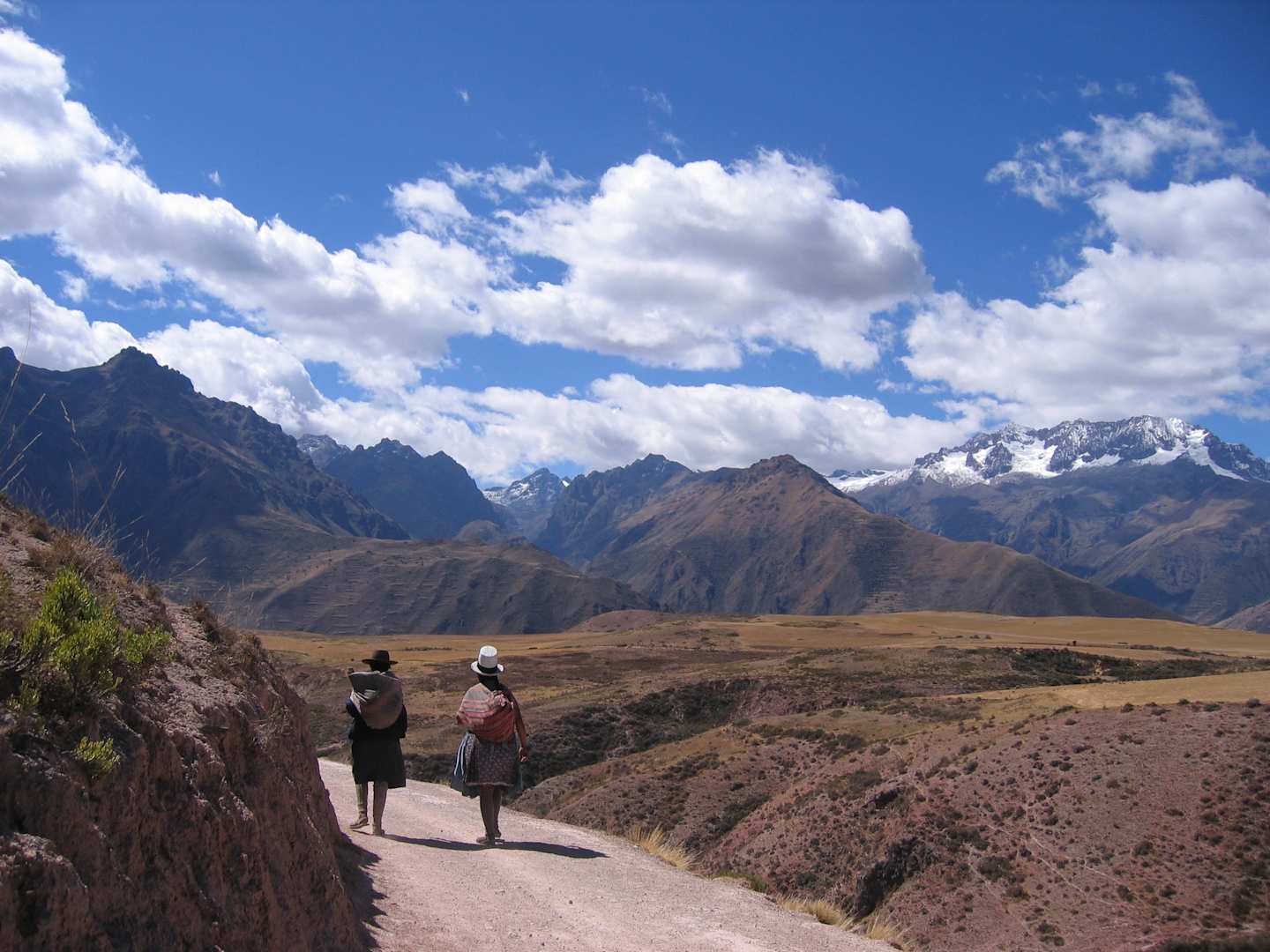 Two people walking on a path in a mountainous landscape with snow-capped peaks in the background and a cloudy blue sky overhead.
