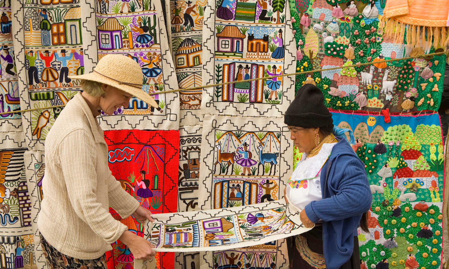 The image depicts a vibrant and colorful marketplace, with a person in a straw hat examining a display of intricate and patterned textiles or artwork in the foreground, surrounded by a lively and visually striking background of various decorative items and structures.