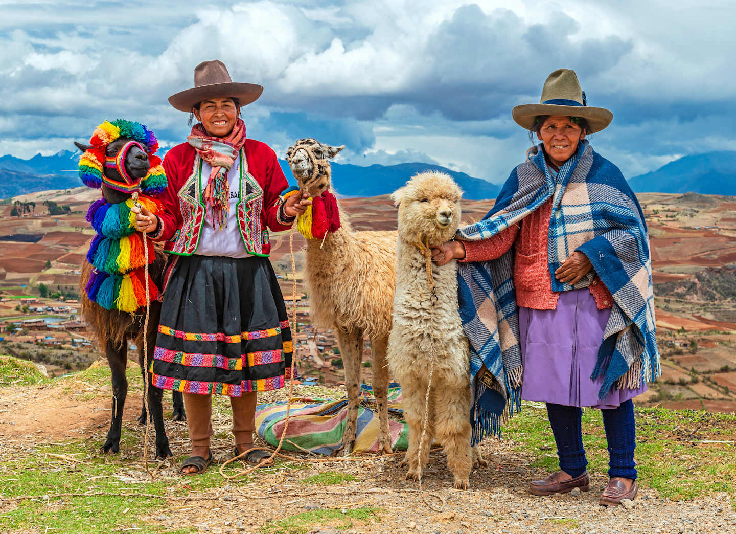 Two individuals wearing traditional Peruvian attire, including colorful ponchos and hats, stand in a scenic mountainous landscape with a llama by their side.