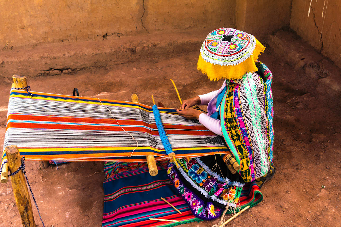 A colorfully dressed person is weaving a vibrant, patterned textile on a traditional loom against a rustic, earthen backdrop.