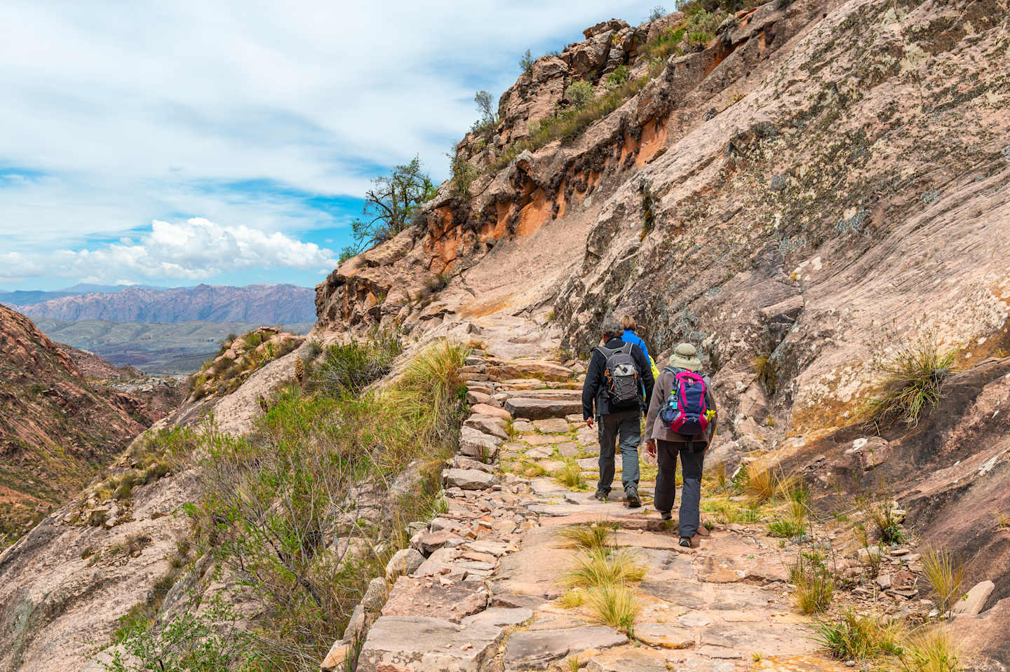 A rugged mountain trail winds through a rocky landscape, with two hikers in the foreground navigating the path amidst the dramatic terrain.