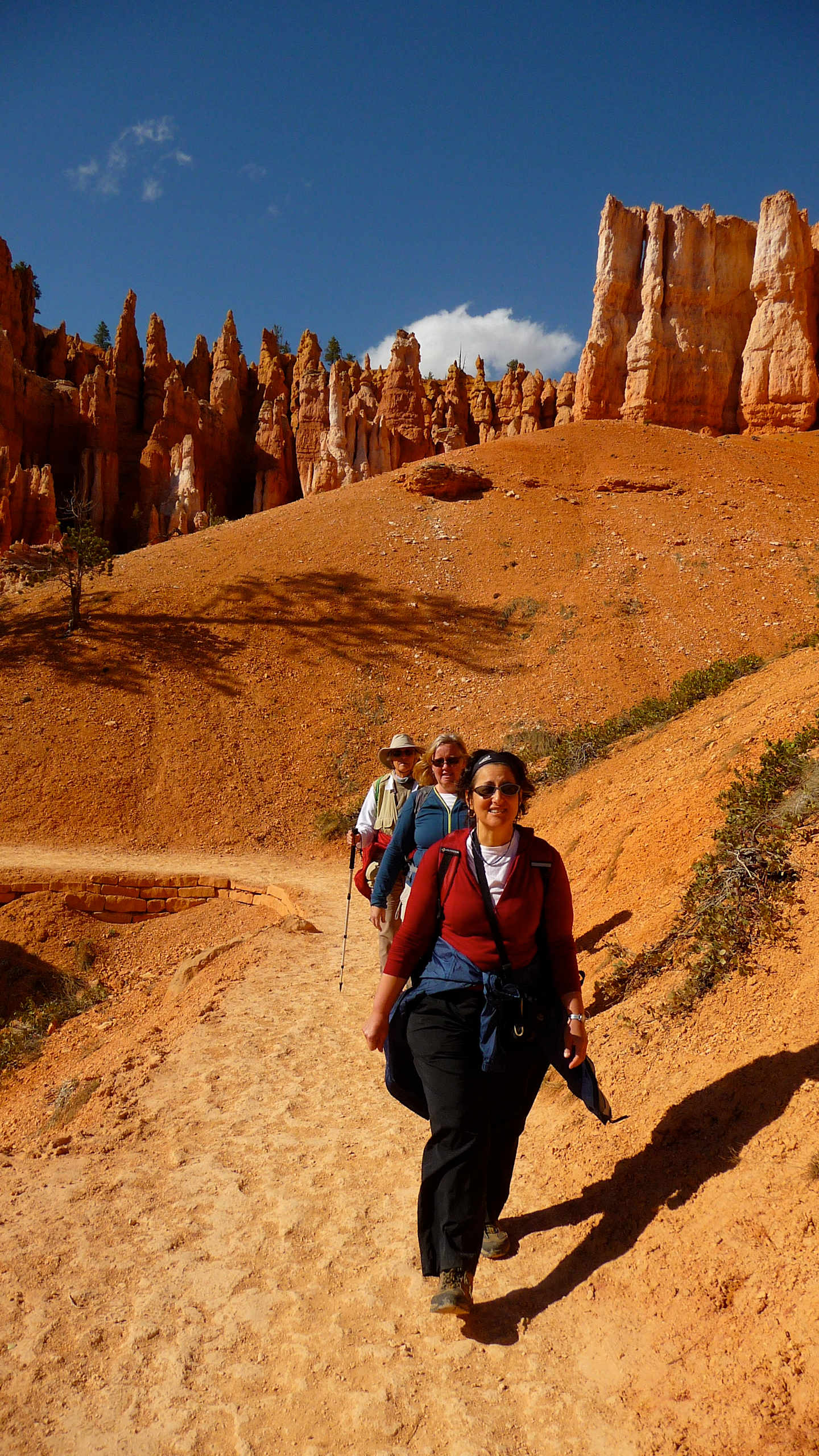 The image shows a group of hikers trekking through a rugged, arid landscape with towering red rock formations in the background, under a bright blue sky with scattered clouds.